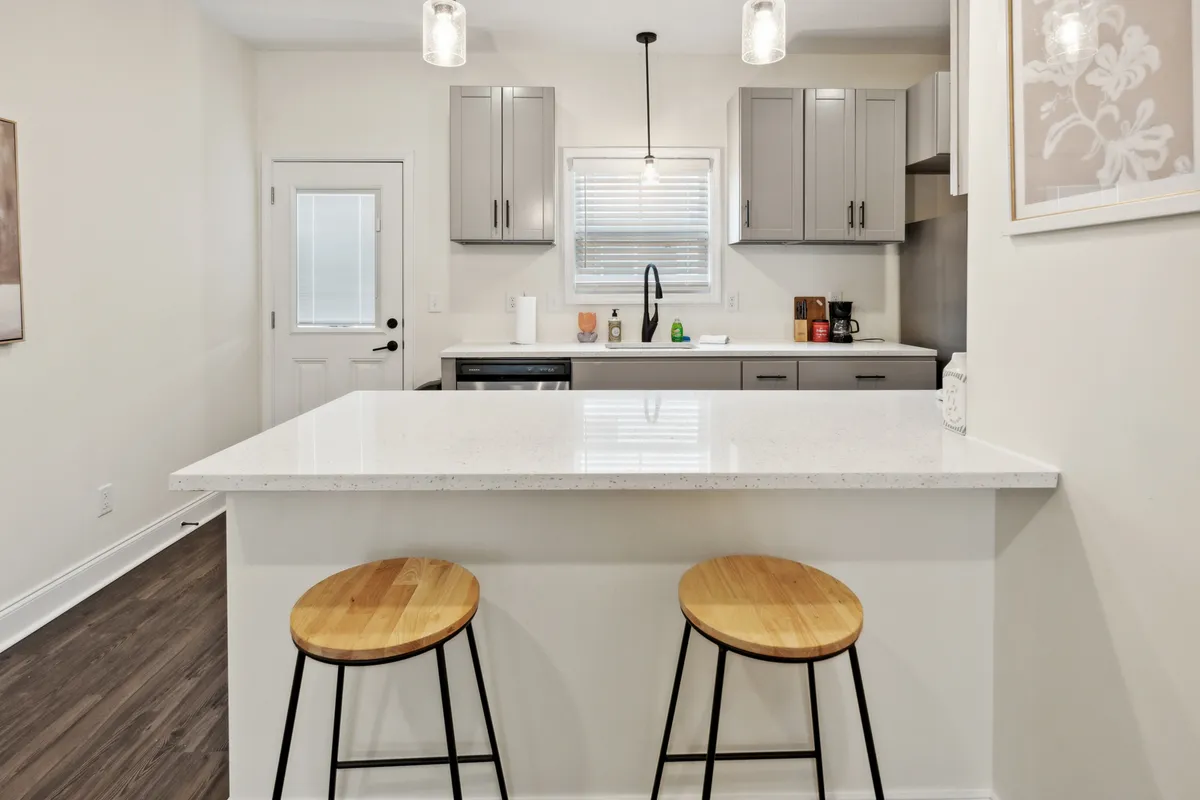 This image showcases a modern kitchen with a central island, bar stools, ample countertop surface, stylish light fixtures, and a sunlit window. It captures people engaged in activities such as cooking, socializing, and enjoying breakfast, highlighting the kitchen's function as a hub for both daily routines and gatherings.