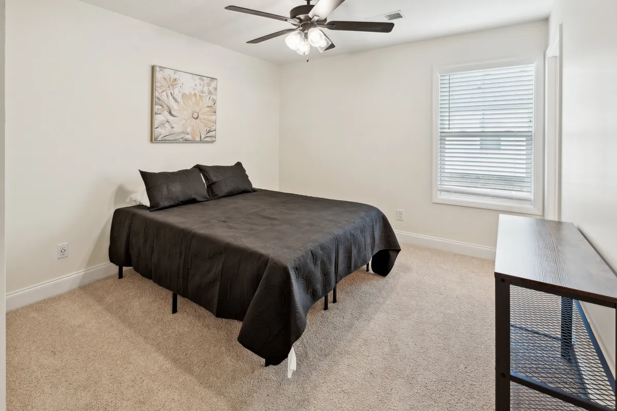 A cozy bedroom featuring a bed with black bedding, floral wall art, a window with blinds allowing natural light, and a nightstand. The atmosphere is peaceful and restful, ideal for unwinding after a busy day.