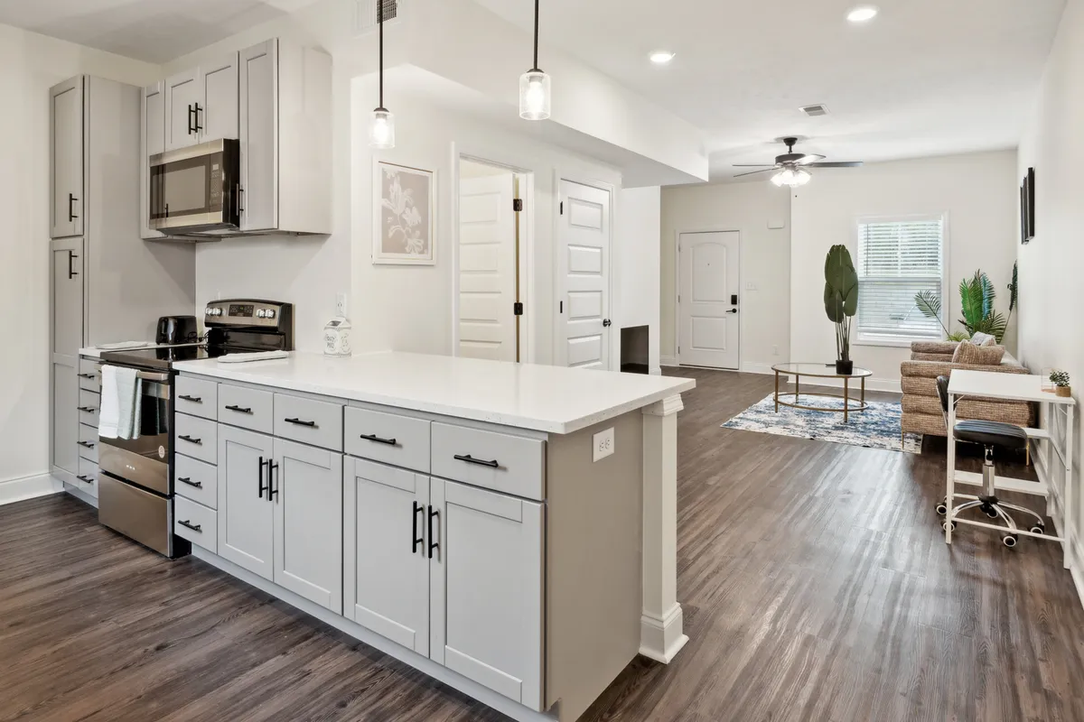 A view that captures a modern kitchen island and stainless steel appliances, with an open sightline to a cozy living area furnished with plush seating. The room is filled with natural light, highlighting the home's airy, interconnected atmosphere.