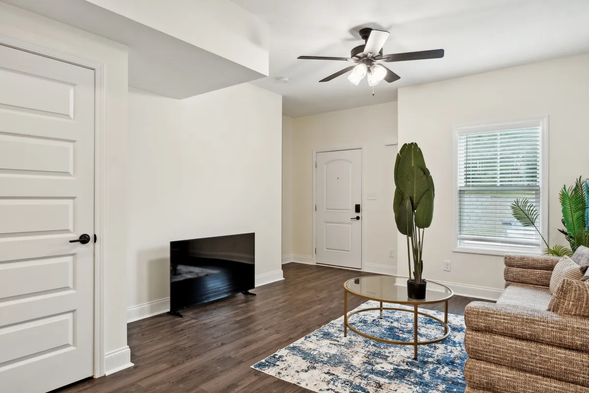 A cozy living room scene featuring a soft sofa, coffee table, potted plant, TV, and a doorway leading into another area. The environment is intimate yet functional, suggesting a space where people can unwind or entertain.